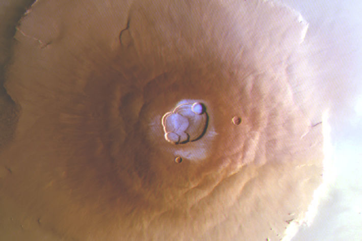 En bild av vulkanen Olympus Mons på Mars tagen ovanifrån. Den brunröda vulkanen breder ut sig över ytan, och i höjden på mitten av vulkanen finns en urholkning som liknar en krater. I denna krater syns vattenis som ett tunt, ljusblått lager som kontrasterar mot den annars enfärgade vulkanen.
