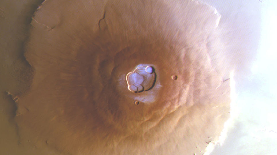 En bild av vulkanen Olympus Mons på Mars tagen ovanifrån. Den brunröda vulkanen breder ut sig över ytan, och i höjden på mitten av vulkanen finns en urholkning som liknar en krater. I denna krater syns vattenis som ett tunt, ljusblått lager som kontrasterar mot den annars enfärgade vulkanen.